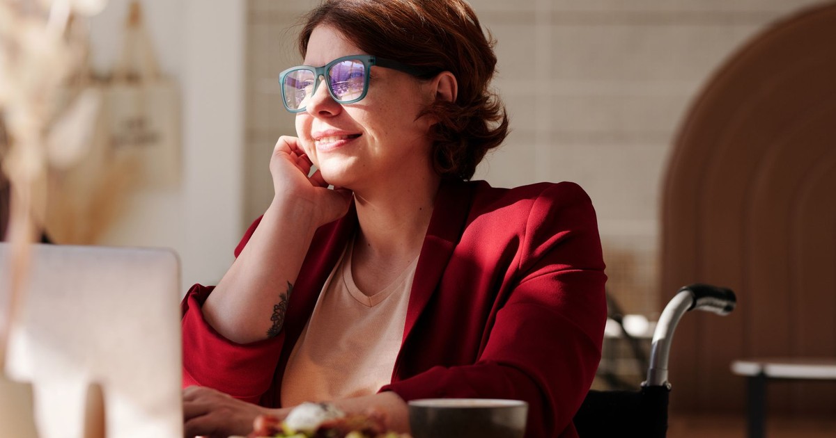 Cheerful woman in a red blazer working remotely on a laptop at a café, enjoying the sunlight.