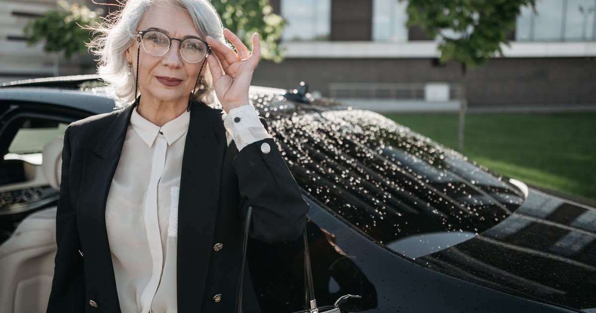 Stylish senior businesswoman with grey hair and eyeglasses standing by a black car outdoors.