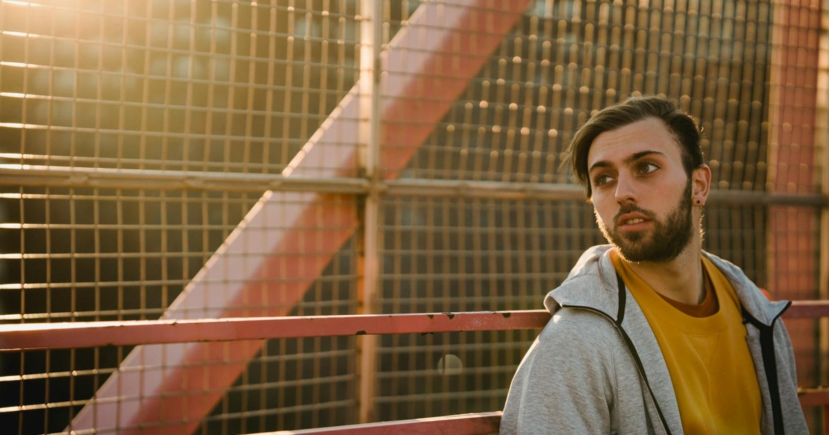 Young contemplative bearded male in casual wear looking away near metal fence in sunlight