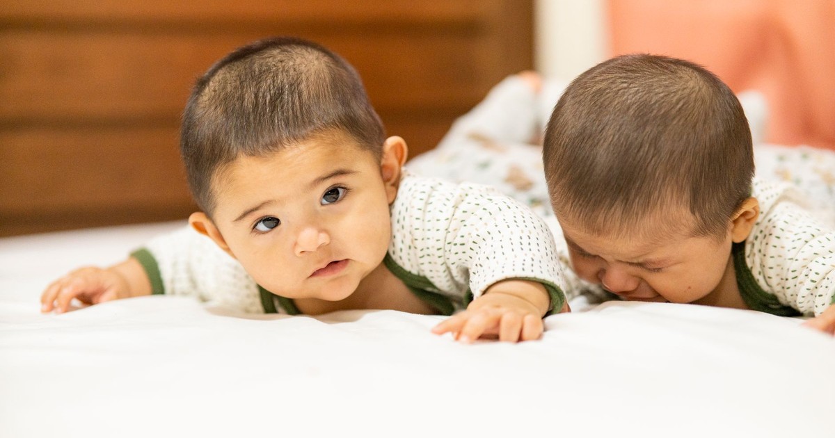 Adorable twin babies in matching outfits lying on a cozy bed indoors, capturing a sweet moment.