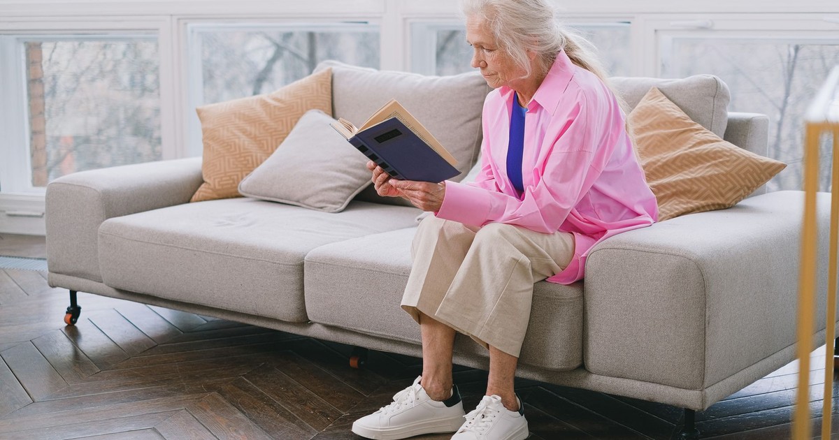 Elderly woman in bright clothing reading a book on a cozy indoor sofa.