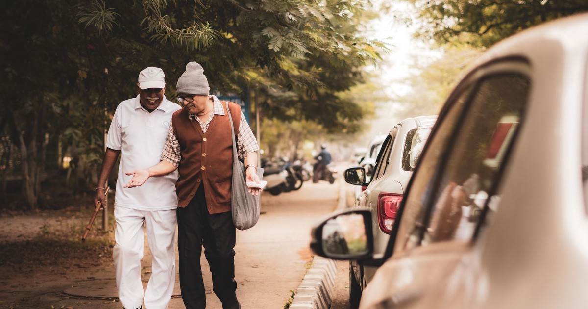 Two elderly men walking arm-in-arm on a tree-lined city sidewalk, sharing a moment.
