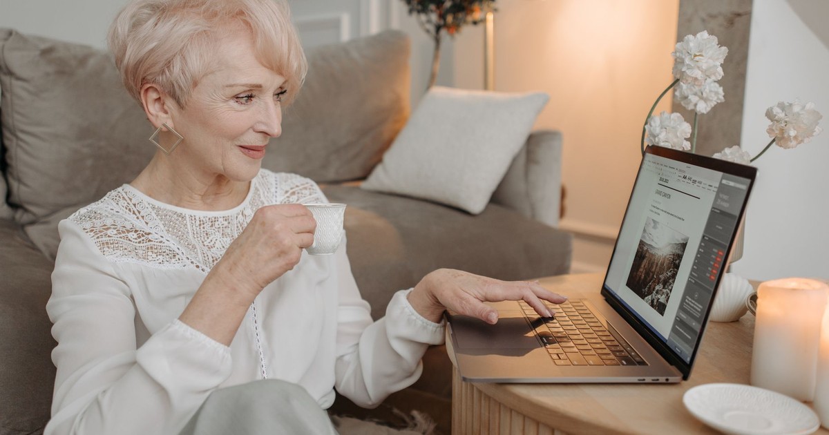Elderly woman with short hair working from home on a laptop, enjoying coffee.