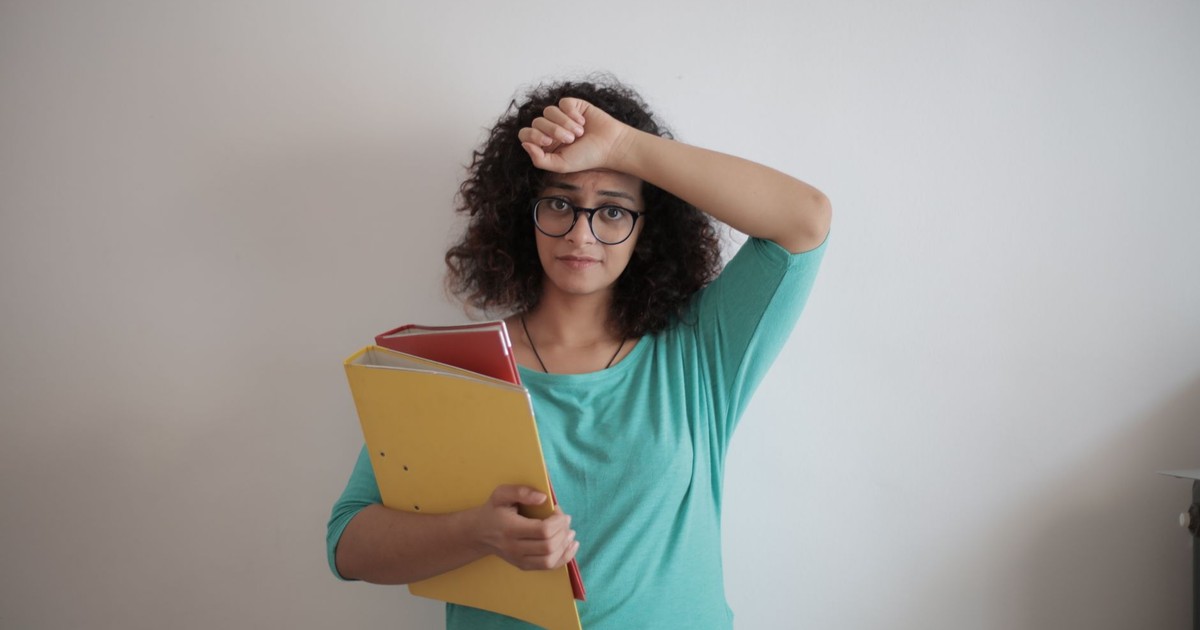 Unhappy overworked female employee in glasses and casual clothes with folders of documents looking at camera with sadness and touching head while standing against white wall in contemporary office