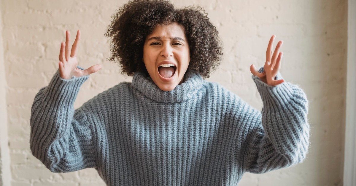 Irritated African American female with raised arms looking at camera while shouting loudly near wall in light room at home