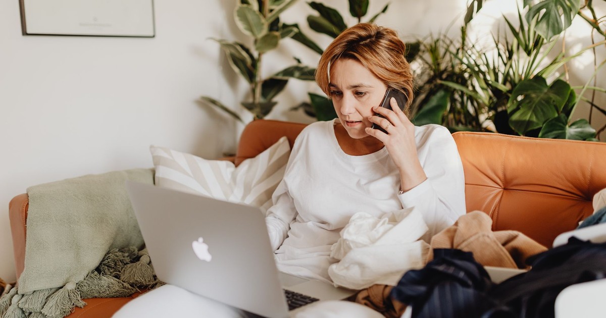 A woman works remotely on a laptop and phone, surrounded by potted plants.