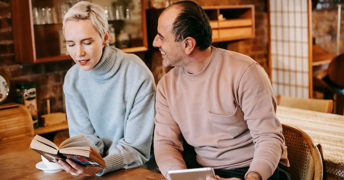 Cheerful middle aged ethnic man with tablet in hands smiling and looking at wife reading book at table while drinking coffee together at home