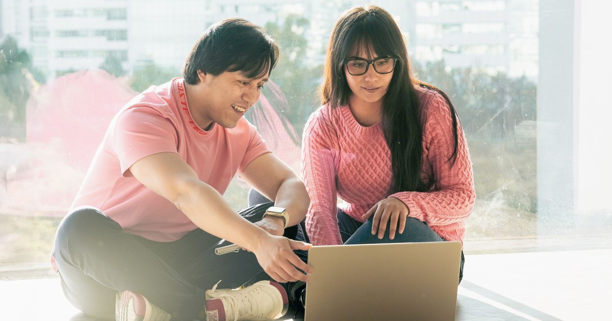 Two young adults in casual attire working together on a laptop by a window.