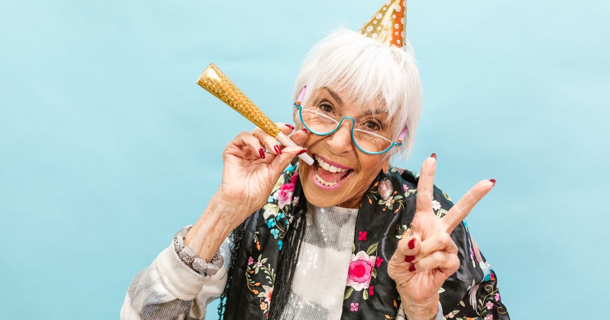 Senior woman in party hat joyfully celebrating, making peace sign on a light blue background.