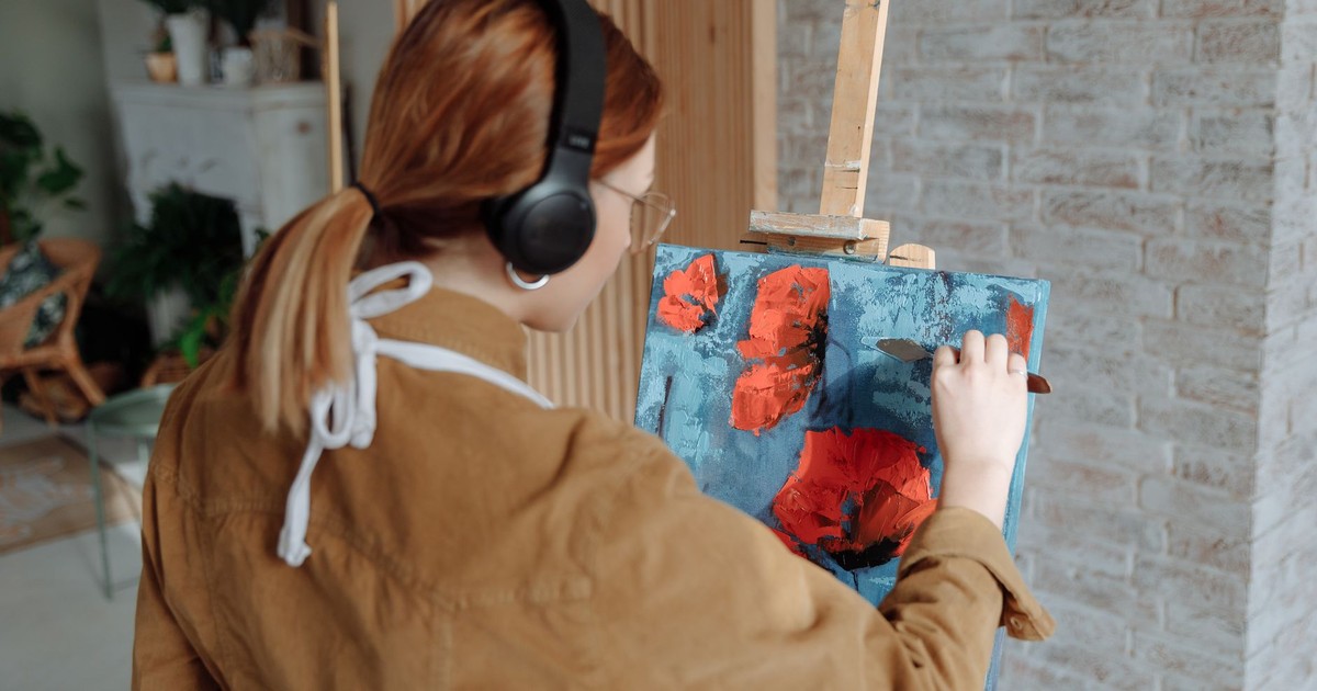 Adult woman using palette knife for painting on canvas with wireless headphones in a cozy studio.