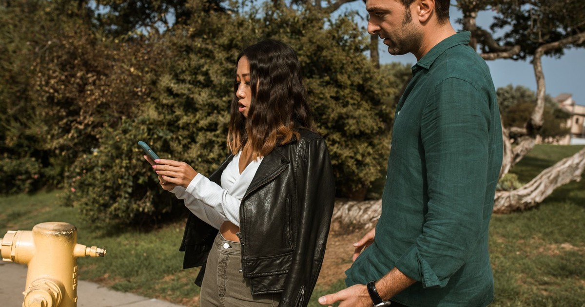 Two adults interacting with a phone, standing outdoors surrounded by trees.