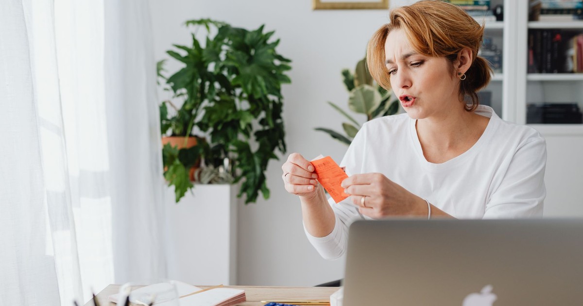Woman reviewing bills at home desk with laptop and plants, managing personal finances.