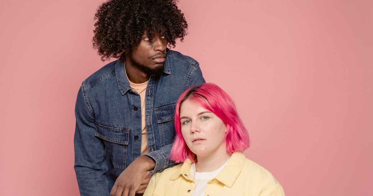 Young black man leaning on shoulder of woman with dyed hair sitting against pink background