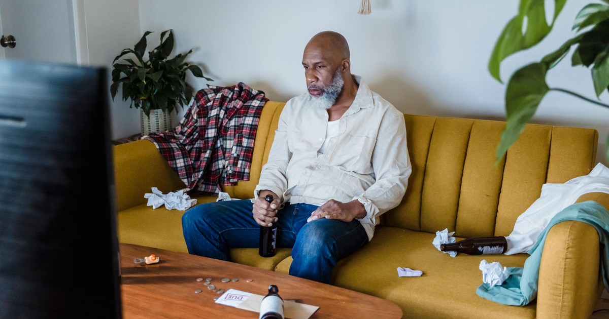 Man sitting on a couch watching TV while holding a beer bottle, surrounded by casual home setting.