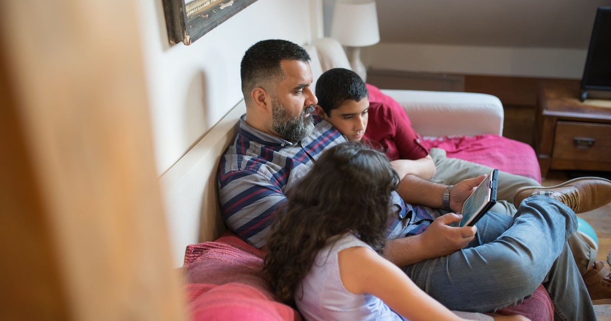 A father and his children relaxing on a couch using a digital tablet in a cozy home setting.