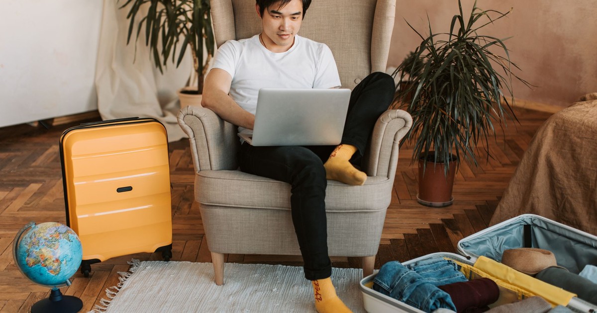 Man sitting on armchair planning travel, using laptop, with suitcases and globe nearby.