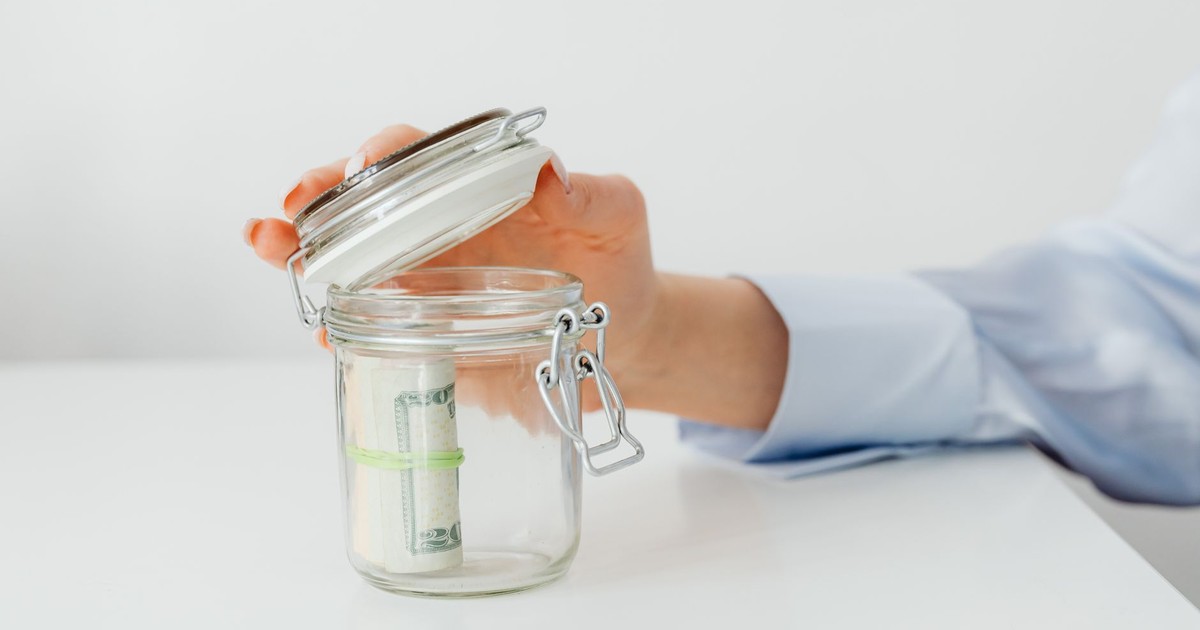 A hand places money in a glass jar on a white table, symbolizing savings.