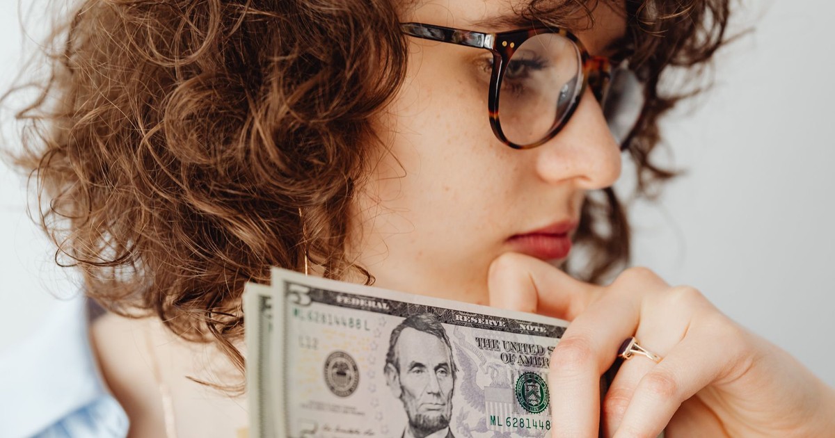 Close-up of a woman with glasses holding US dollar bills. Thoughtful expression.