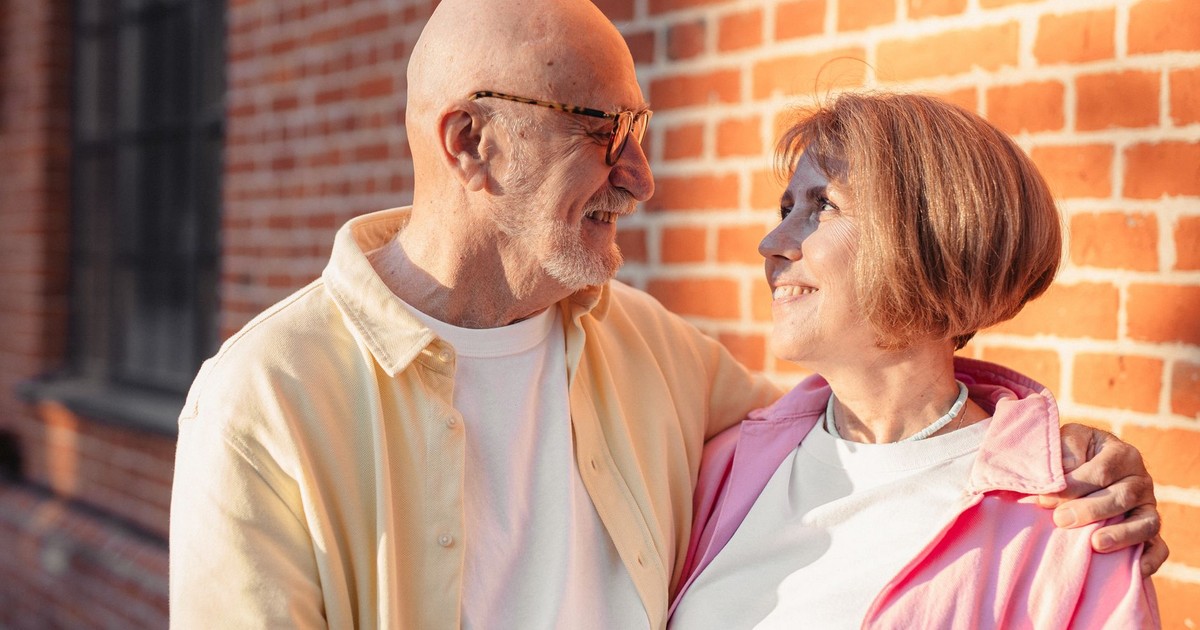 Elderly couple smiling and hugging against a sunlit brick wall, enjoying a moment of love.