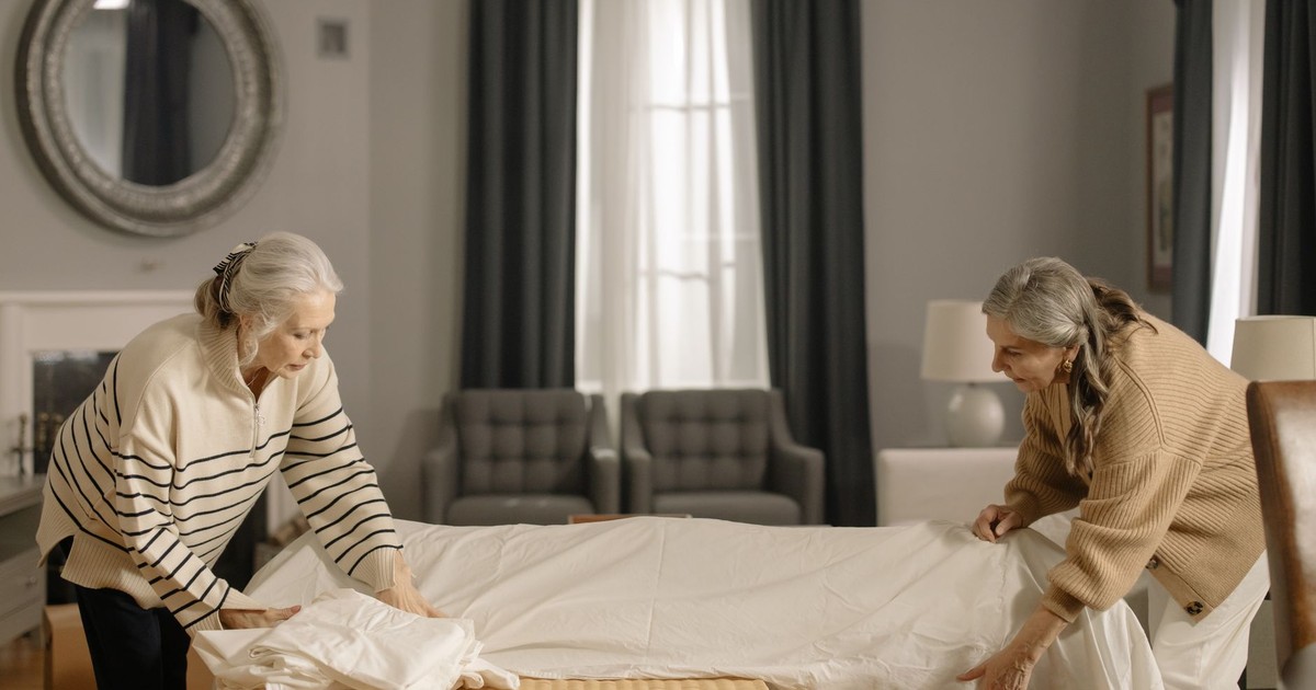 Two senior women folding sheets in a living room, preparing for moving out.