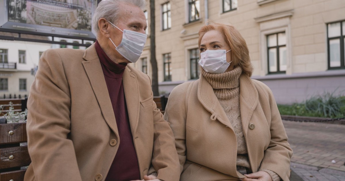 Elderly couple in brown coats wearing masks and sitting outdoors, symbolizing care and romance.