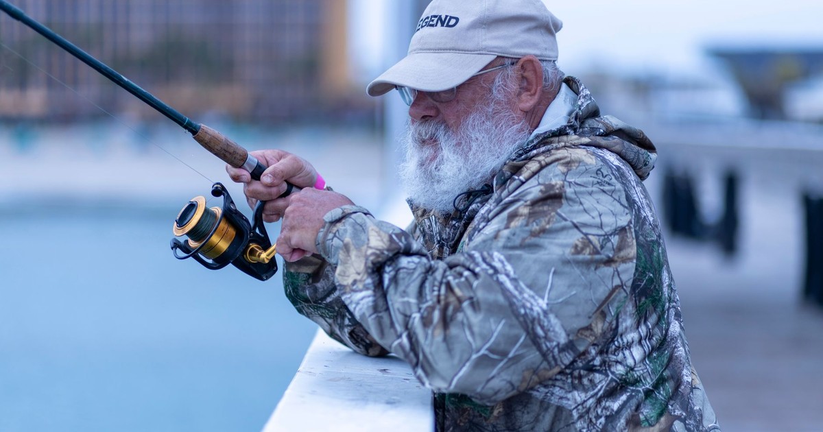 Elderly man with a beard fishing by the pier in a camo jacket on a cloudy day.