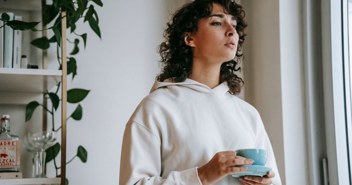Low angle of female in hoodie attentively looking away while standing with cup of coffee