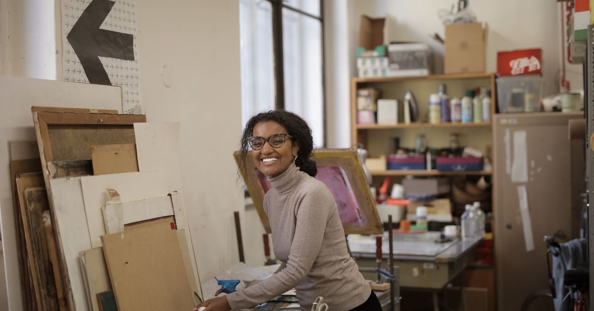 Happy young black female artist in casual clothes and eyeglasses standing in messy art workshop laughing looking at camera on daytime