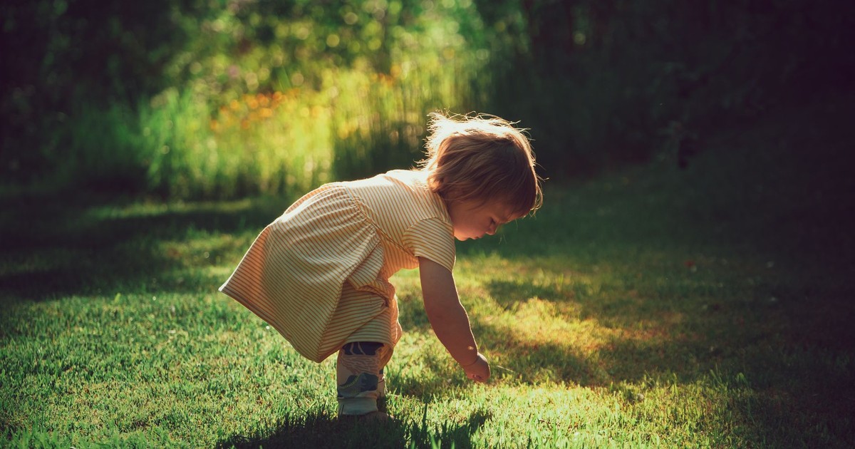 Side view of adorable kid in dress leaning forward on bright green meadow with shade while looking down in back lit