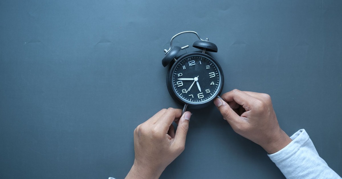 Hands adjusting a classic black alarm clock on a blue background with ample copy space.