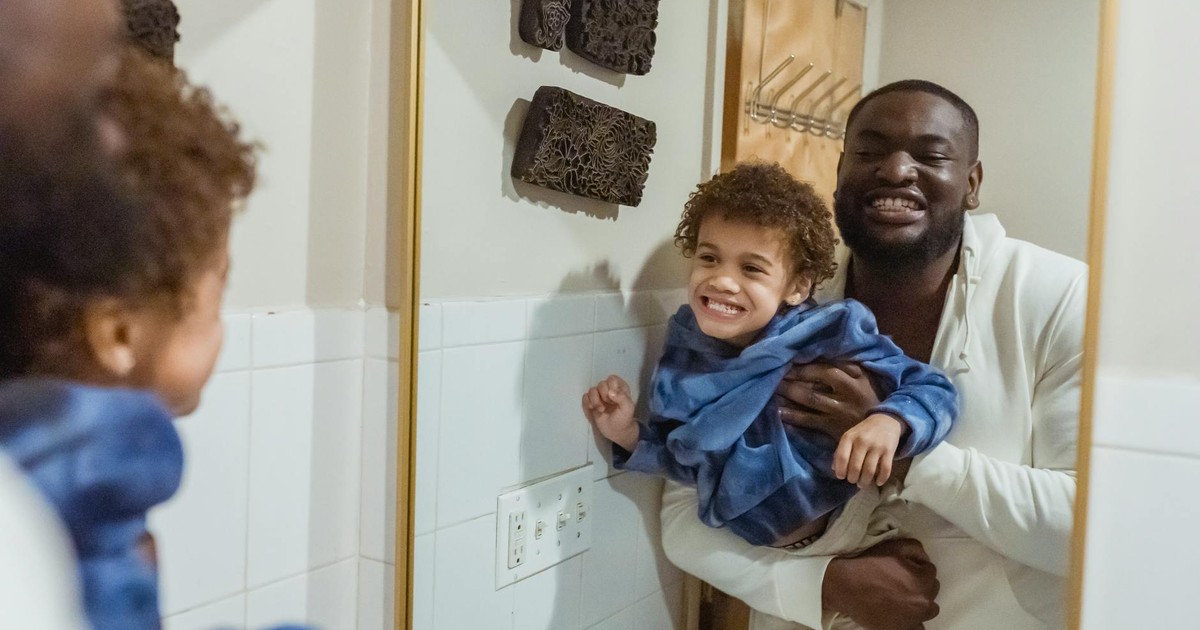 Content African American father with son in hands looking at reflection of mirror and showing teeth while standing in bathroom