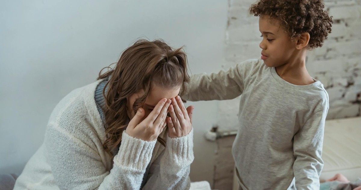 Caring African American son touching shoulder of upset faceless mother covering face while sitting in light room near wall at home