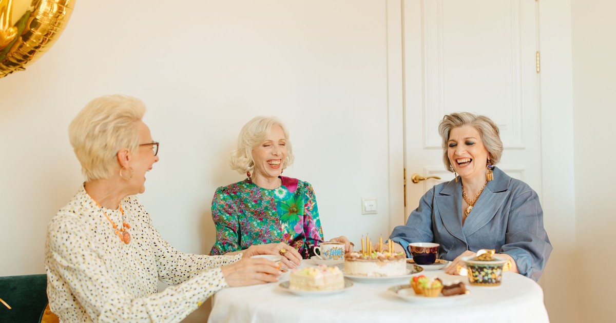 Three senior women laughing and enjoying a tea party with cakes indoors.