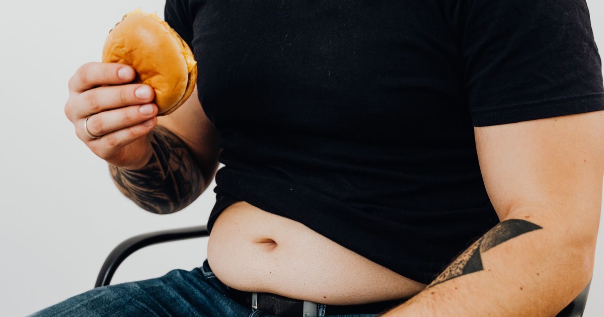 Close-up of a man holding a burger, showcasing his torso and tattooed arm. Casual and relaxed setting.