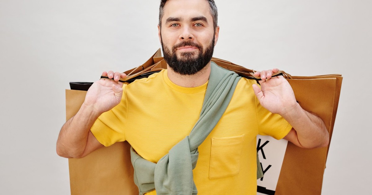 Bearded man in a yellow shirt holding shopping bags against a neutral background.