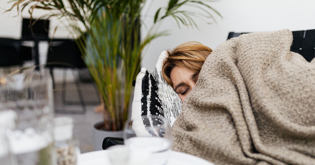 A woman resting on a couch covered by a blanket, indoors with plants.