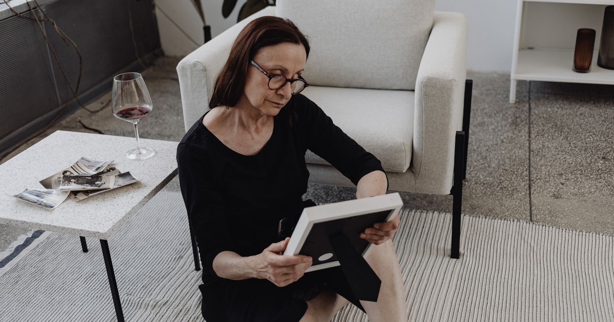 A mature woman sits on the floor in quiet reflection, holding a picture frame, a glass of wine nearby.