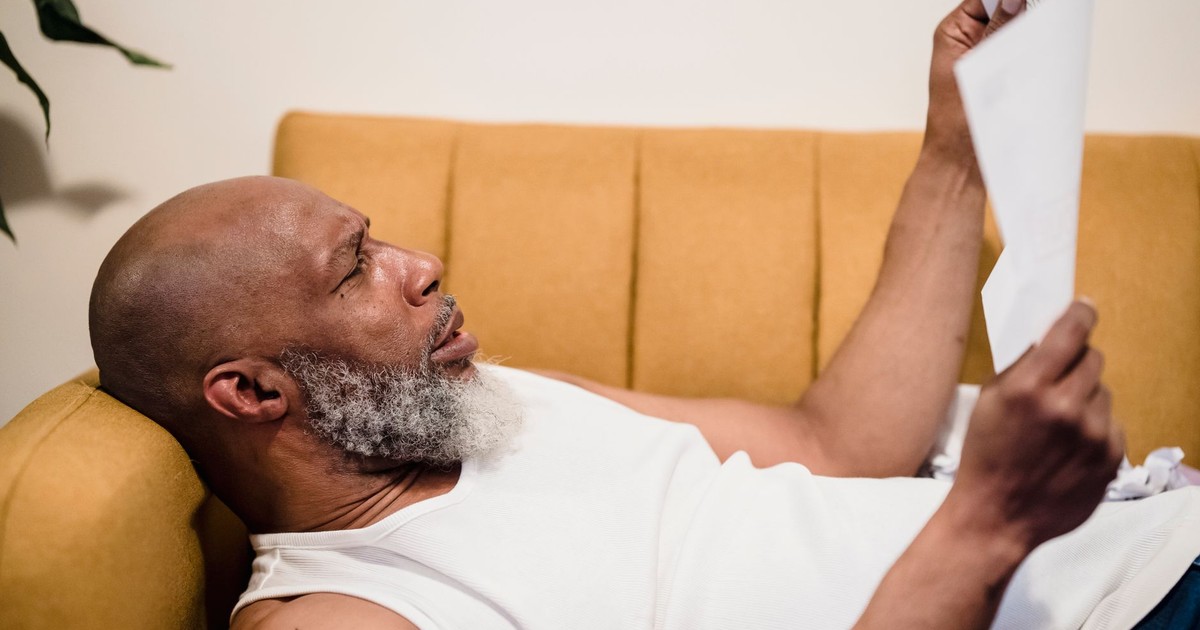 Bald man with facial hair lies on a mustard couch, reading documents in a relaxed setting.