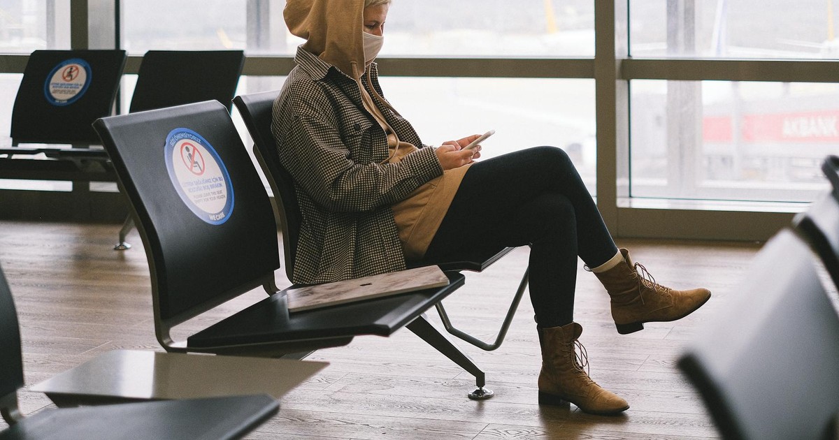 A woman in a hoodie sits in an airport lounge, practicing social distancing while using her phone.