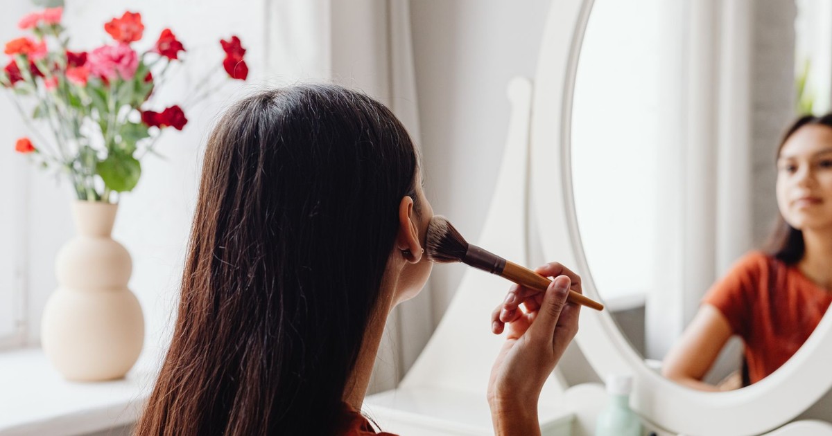 A woman applying makeup with a brush while facing a mirror indoors.