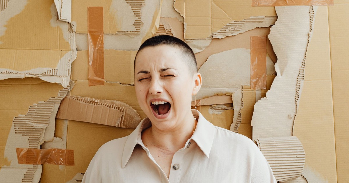 A woman expressing frustration and stress, screaming against a torn cardboard backdrop indoors.