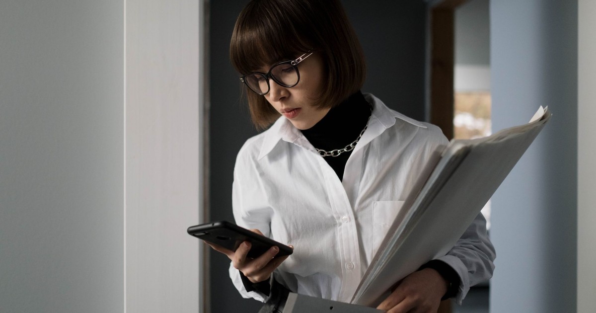 Young woman with eyeglasses, holding documents, checking smartphone indoors.