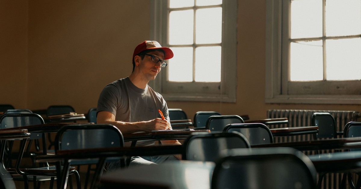 A university student in a cap studying alone in an empty classroom, lit by natural sunlight.