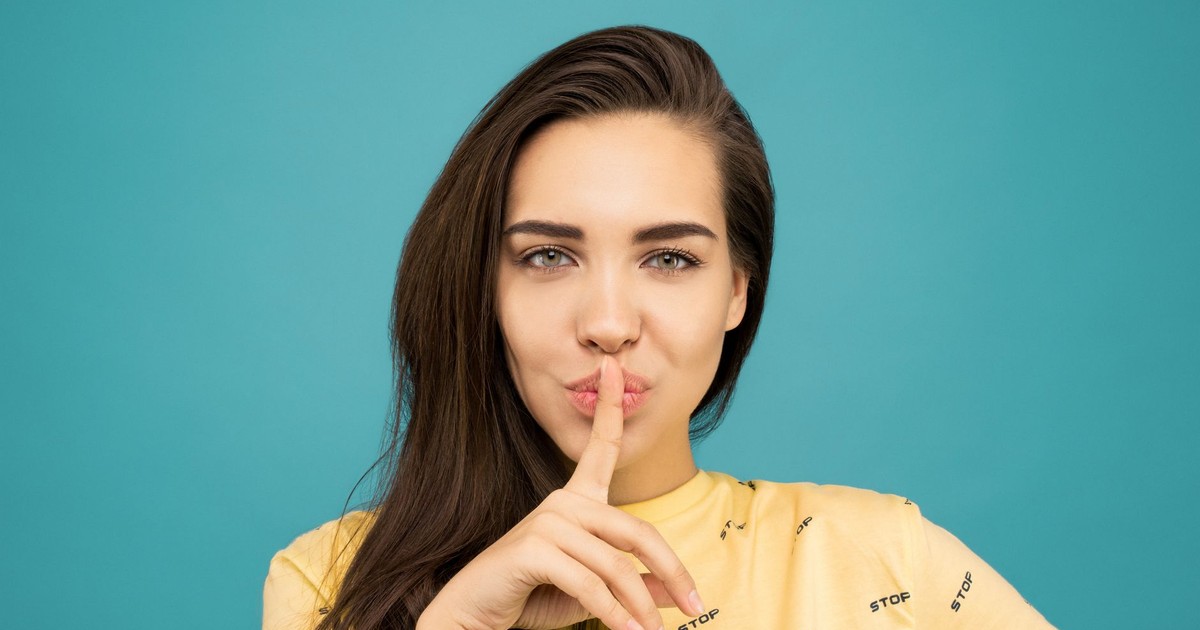 Portrait of a woman with brown hair posing with a hush gesture against a blue background.