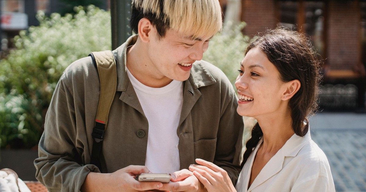Romantic young ethnic lady with long dark hair smiling and touching hand of Asian boyfriend using smartphone on city street