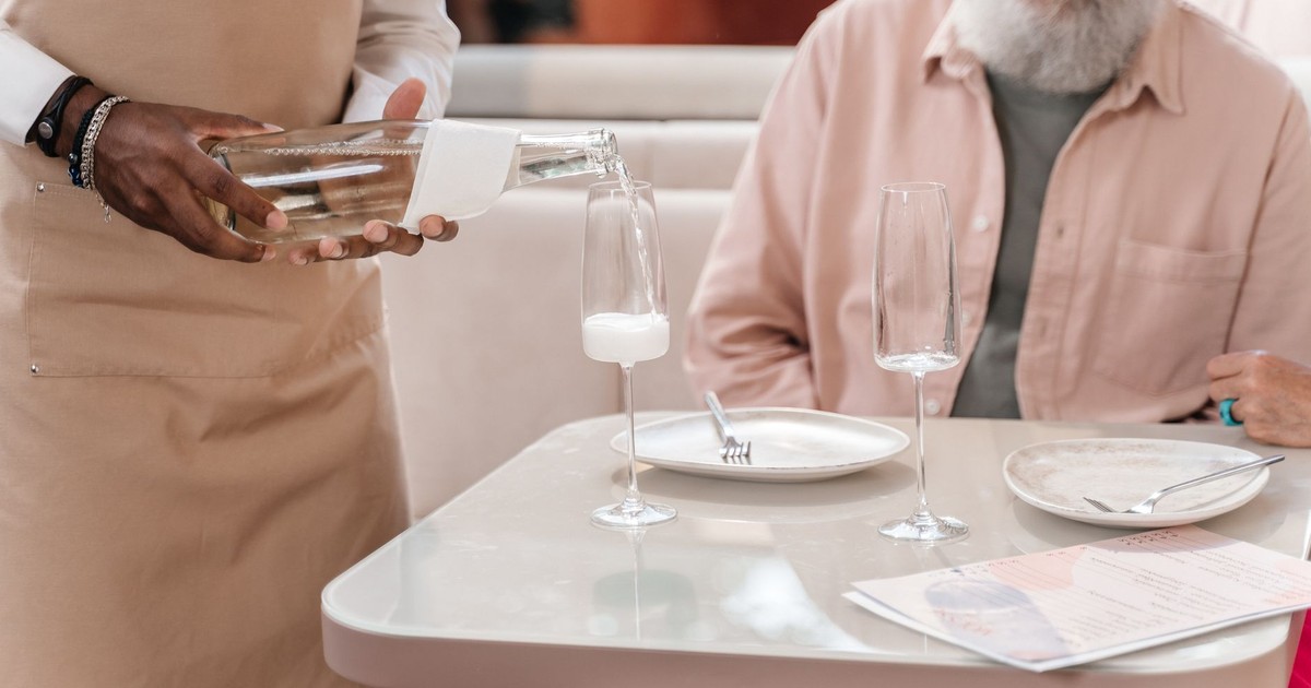 A waiter pouring a drink for an elderly man seated at a restaurant table. Focus on hospitality.