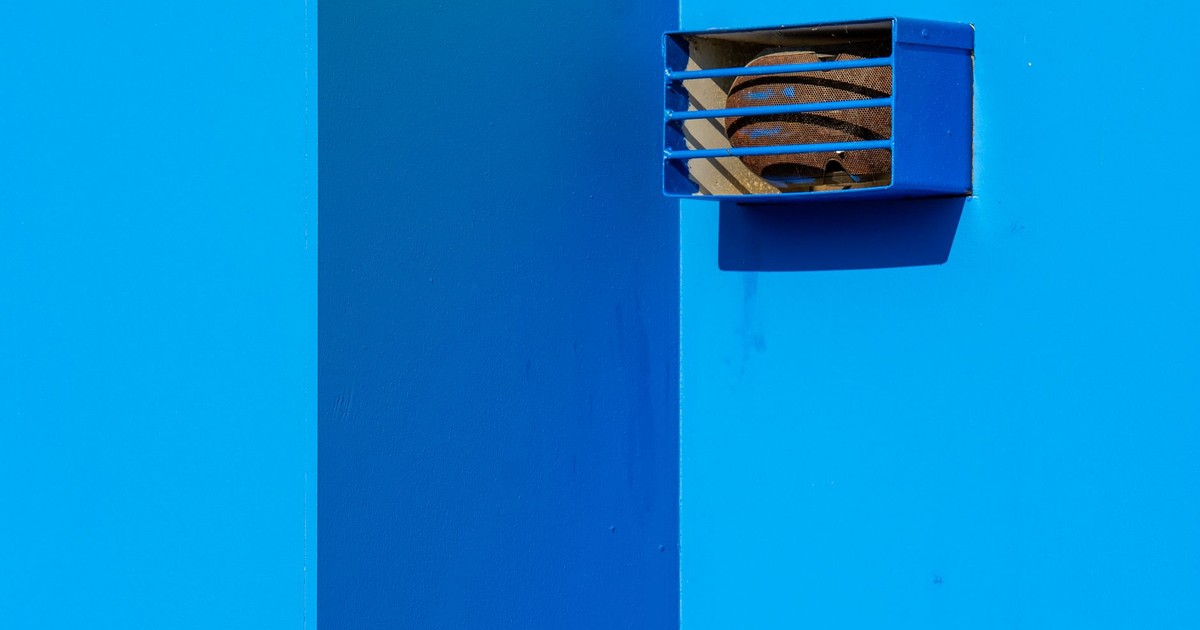 A minimalist shot of a blue wall with rusted vent on sunny day highlighting geometric simplicity.