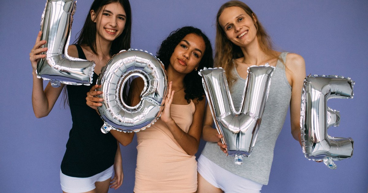 Three diverse women holding silver love balloons against a purple backdrop. Friendship and celebration theme.