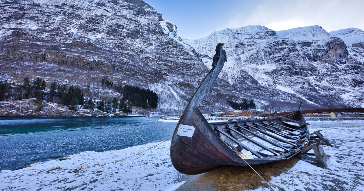 A wooden Viking ship rests on the snowy shores of Aurlandsfjord, Norway, with rocky mountains in the background.