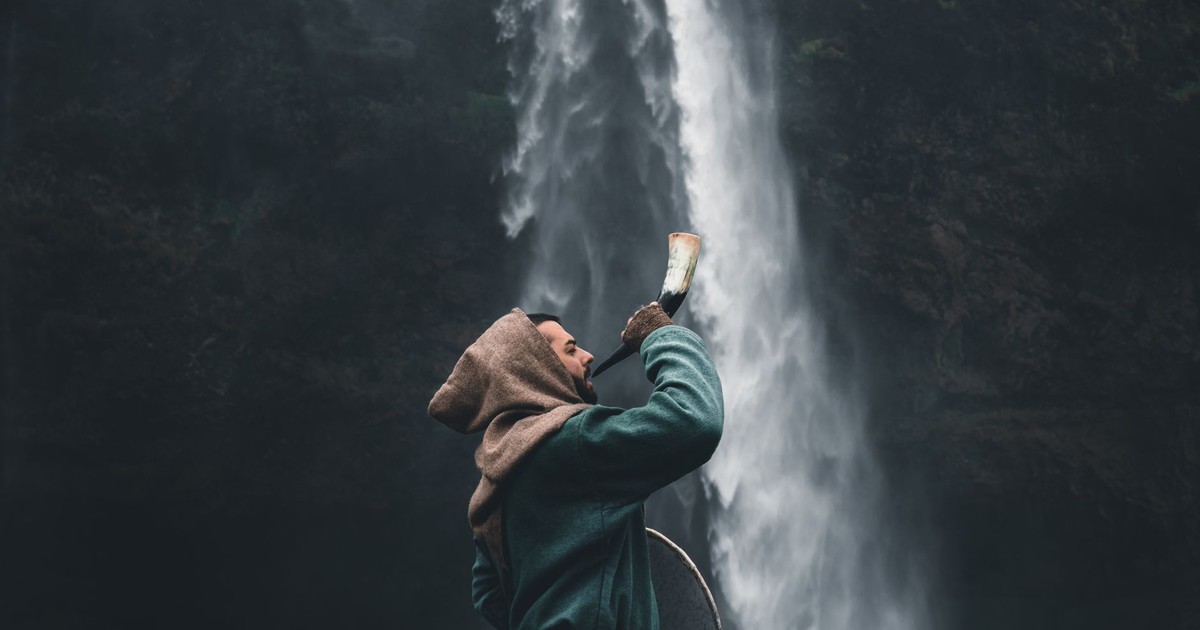 A warrior in a hooded cloak standing by a waterfall in Iceland, holding a horn.